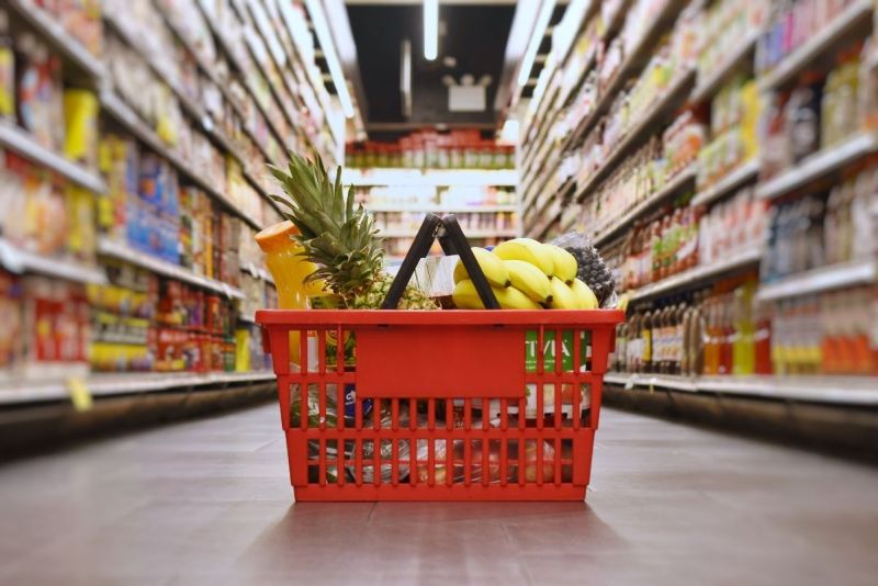 Red shopping basket filled with fresh fruits and vegetables in grocery store aisle.