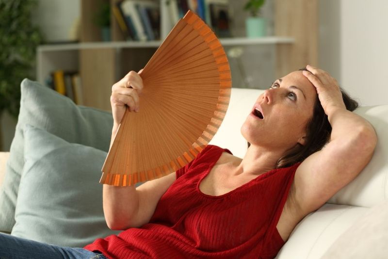 Woman in red top sitting on a couch fanning herself while experiencing a hot flash.