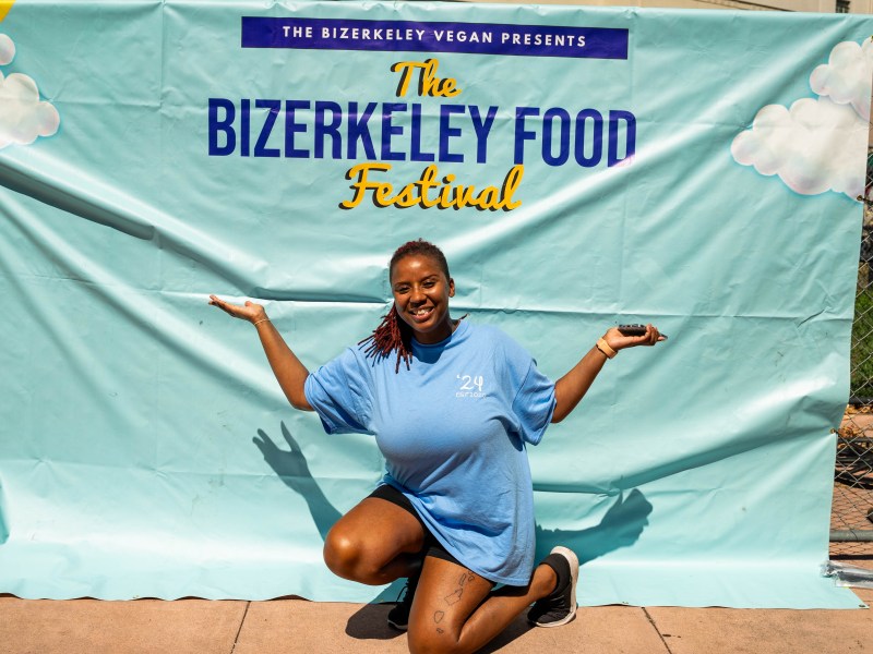 Erika Hazel stands in front of The Bizerkeley Food Festival sign, an aqua blue with orange and navy writing. Erika Hazel kneels down, wearing a light blue festival shirt with her arms in the air.