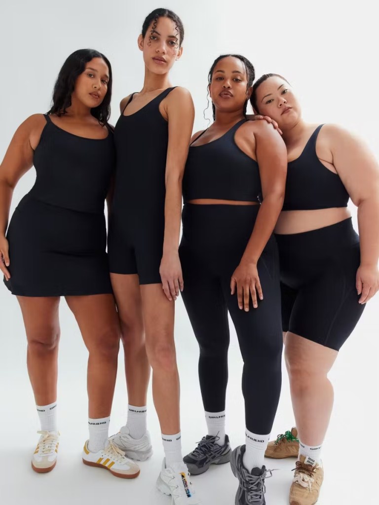 Four diverse women wearing matching black Girlfriend Collective activewear pose confidently against a white background. Each model showcases a different fit and body type, celebrating size inclusivity in sustainable fashion.