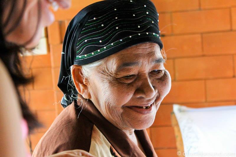 An older Thai woman grins. On her head is a black head covering with tiny blue dots and larger blue dots on the bottom. She is wearing a brown shirt. Behind her is a wall of orange bricks