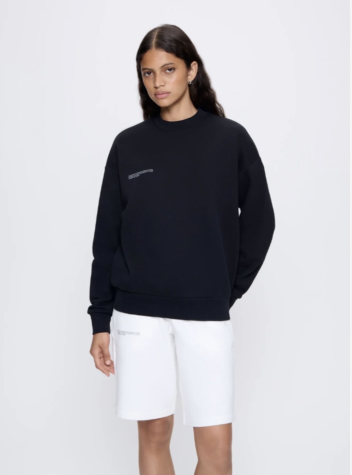A model in a black PANGAIA crewneck sweatshirt and white shorts stands confidently with a neutral background.