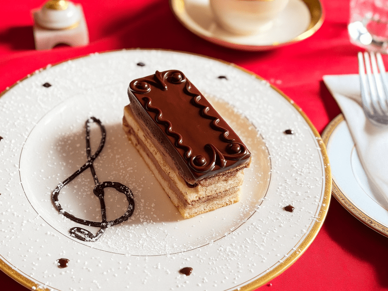 A rectangular slice of vegan opera cake topped with glossy chocolate ganache and intricate decorative swirls, served on an elegant white plate with gold trim. The plate is dusted with powdered sugar and features a chocolate treble clef design beside the dessert.